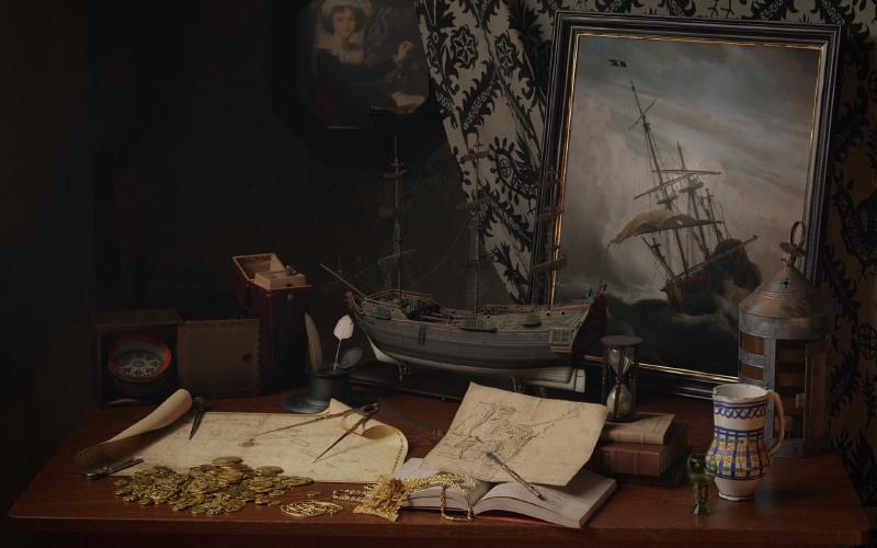 A 3D scene depicting a still life with numerous nautical objects (maps, compass) related to navigation, as well as a painting of a ship in a storm, gold coins and Mesoamerican gold artifacts.